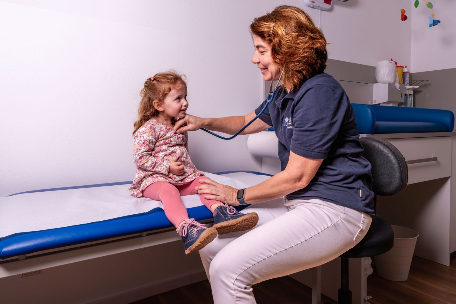 Frau Dr. med. Sandra Dermatidis Fachärztin für Kinder- und Jugendmedizin behandelt ein kleines Mädchen, das auf der Behandlungsliege sitzt. Wir gerade mit einem Stethoskop abgehört. Beide lächeln.